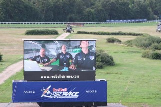 The large screen showing Team #99 cheering on Michael from the hangar.